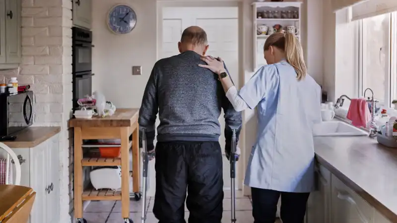 Senior wird von einer Helferin zu Hause beim Laufen mit Krücken unterstützt